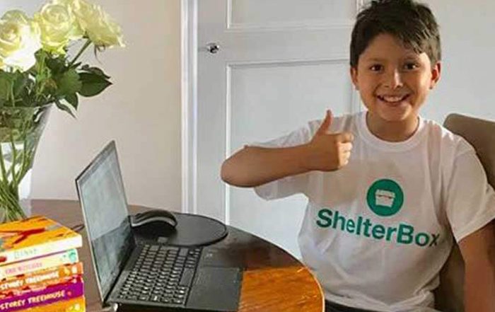 Boy wearing a ShelterBox tshirt sitting at a table with a laptop and pile of books on it