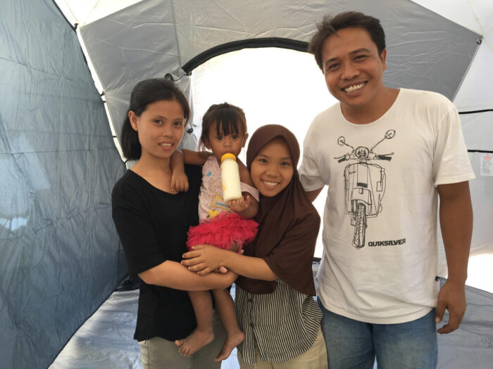 An Indonesian family inside a ShelterBox tent. Young child drinking from a milk bottle being held by her mother.