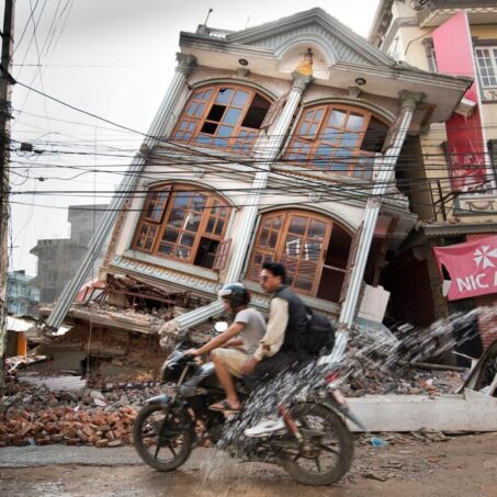 two young men on a motorbike passing a leaning devastated house