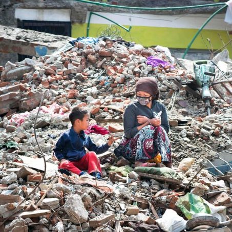 A lady wearing a mask sitting on rubble and debris with a young boy in Nepal