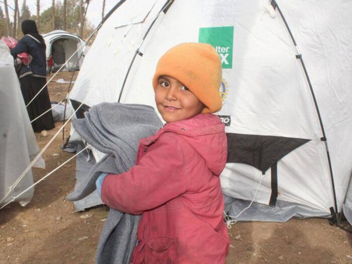 Young Syrian child holding grey blankets with an orange hat standing in front of a ShelterBox tent