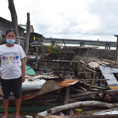 woman in facemask stands next to destroyed home in philippines