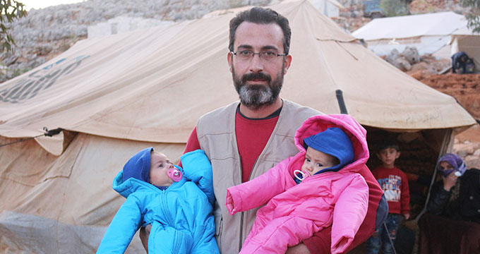 man holding two babies wearing winter clothes in a Syrian camp