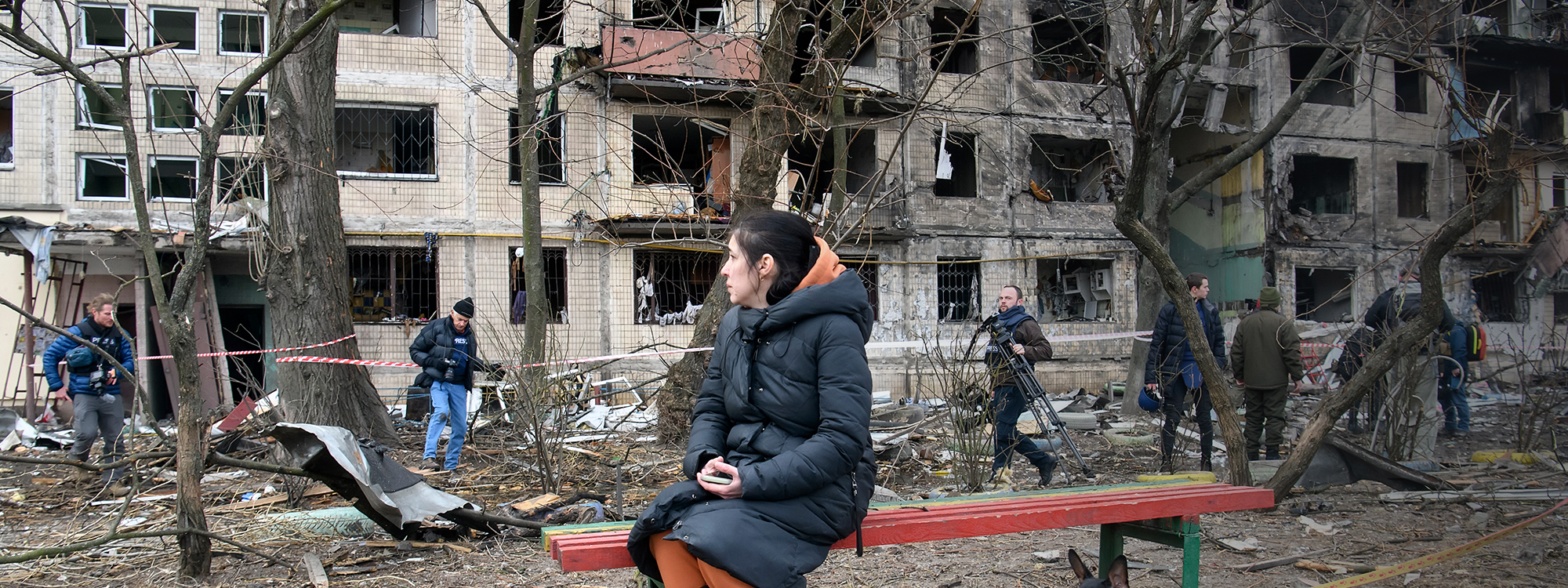 Woman sits on a bench with destroyed buildings behind her