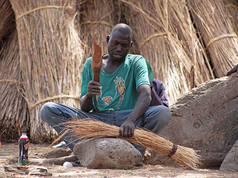Nigerian man sits on the ground making a sweeping brush from dried grasses