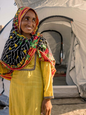 Woman in yellow smiles outside emergency relief tent in Pakistan after flooding