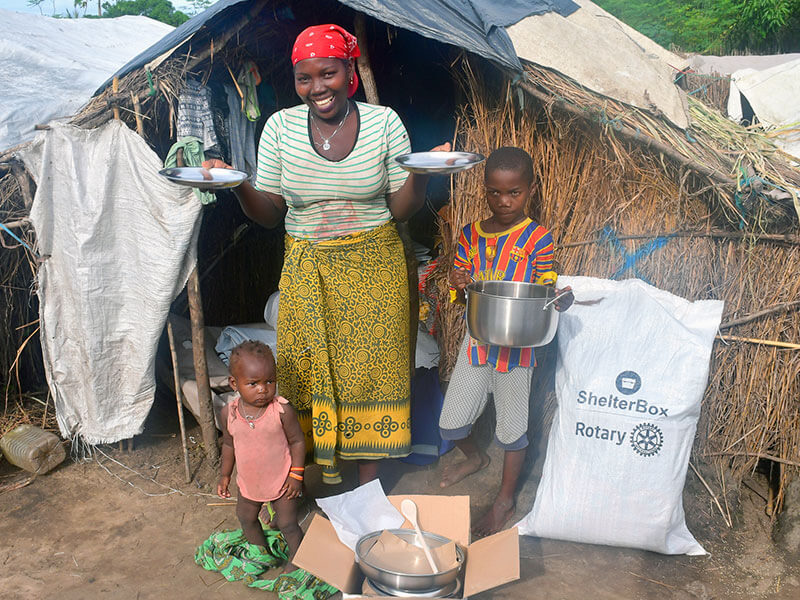 Smiling woman stands outside grass hut holding kitchen items in Mozambique