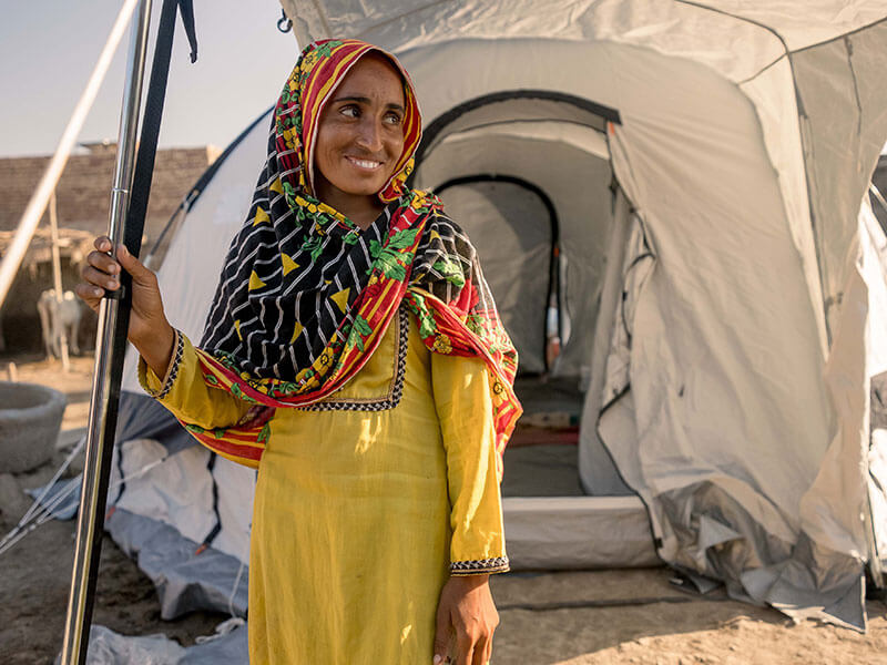Woman in yellow smiles outside emergency relief tent in Pakistan after flooding