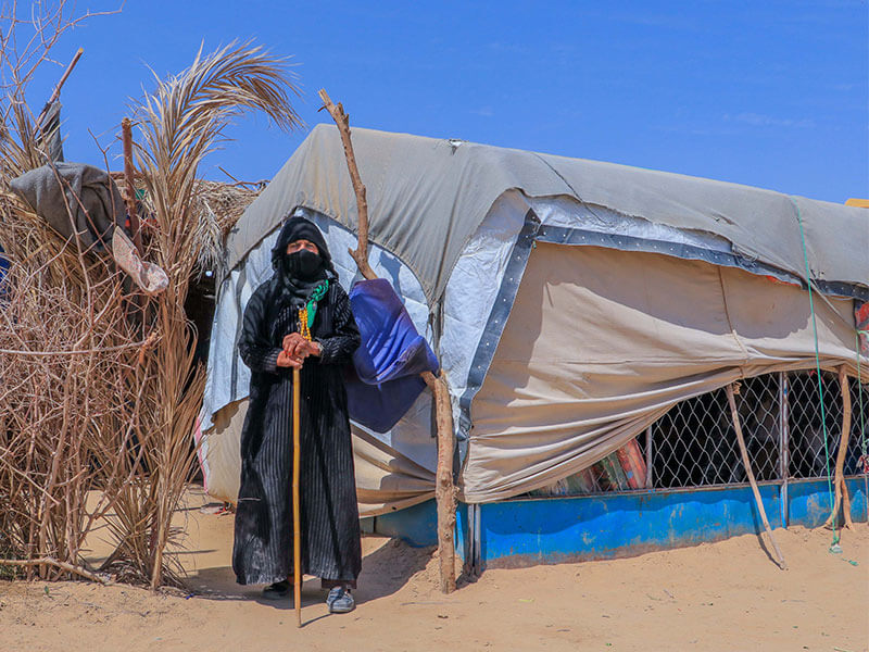 Yemeni woman with walking stick stands outside her shelter