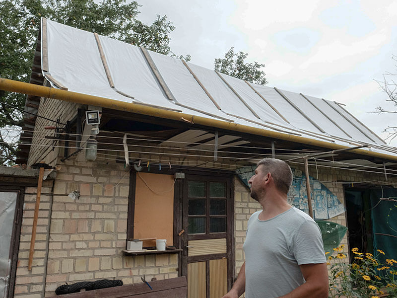 Ukrainian man in grey tshirt looks back at his house, the roof has been repaired with tarpaulin after bombing