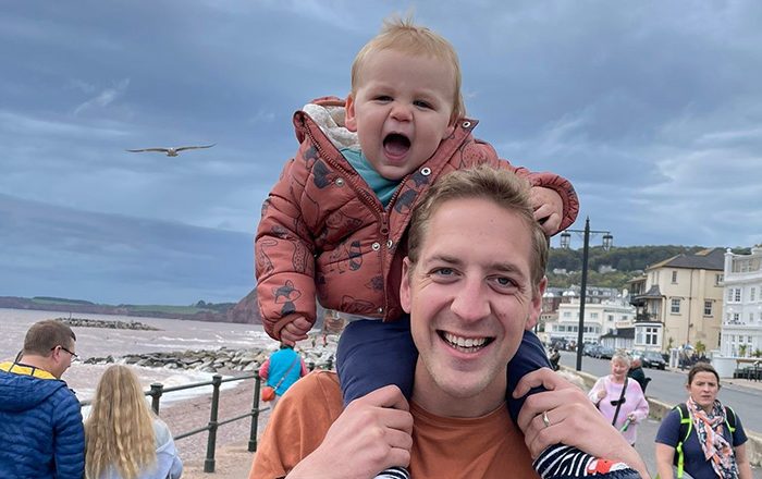 Man holding baby on his shoulders next to a beach