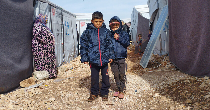 two young boys standing in the middle of a displacement camp, wearing winter coats