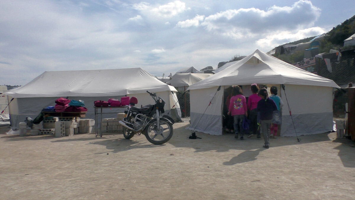 Two large white tents pitched on a sandy floor with children and a motorbike around