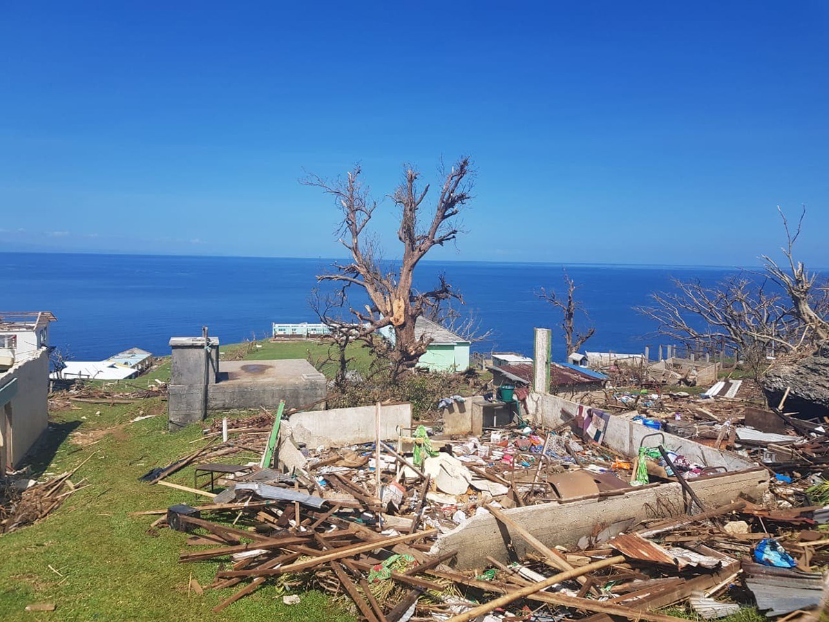 Debris from destroyed homes on the ground next to a tree, with a backdrop of blue sky and the ocean