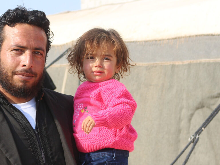 Man holding a child next to a tent in Syria