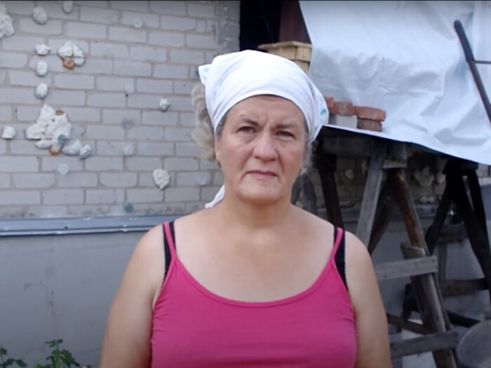 Woman standing in front of a damaged house in Ukraine, part of which is covered in a tarpaulin