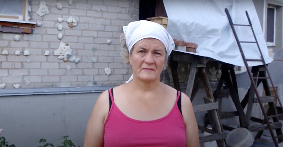 Woman standing in front of a damaged house in Ukraine, part of which is covered in a tarpaulin