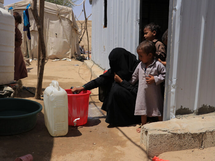 Woman filling up a water bottle watched by two children outside a shelter in Yemen