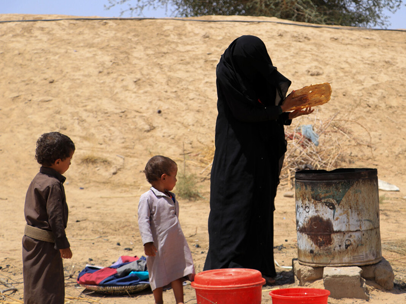 Woman placing materials into a metal barrel next to two small children in Yemen
