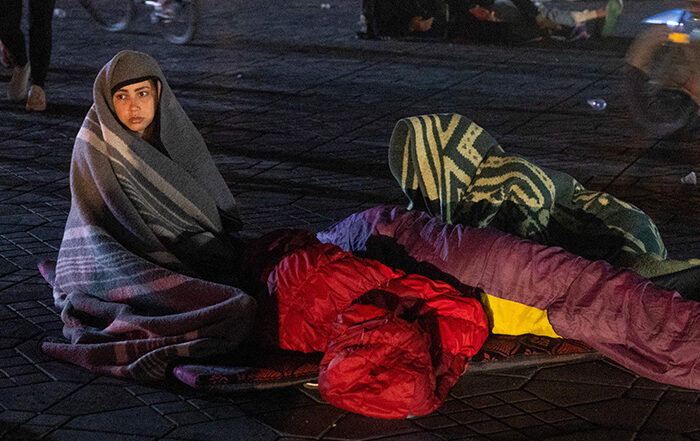 Woman sitting outside in a street at nighttime with sleeping bags around after an earthquake in Morocco.