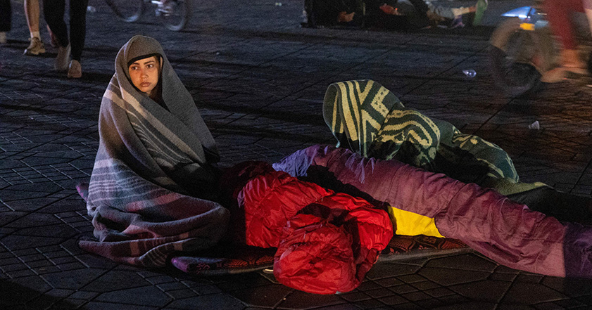 Woman sitting outside in a street at nighttime with sleeping bags around after an earthquake in Morocco.