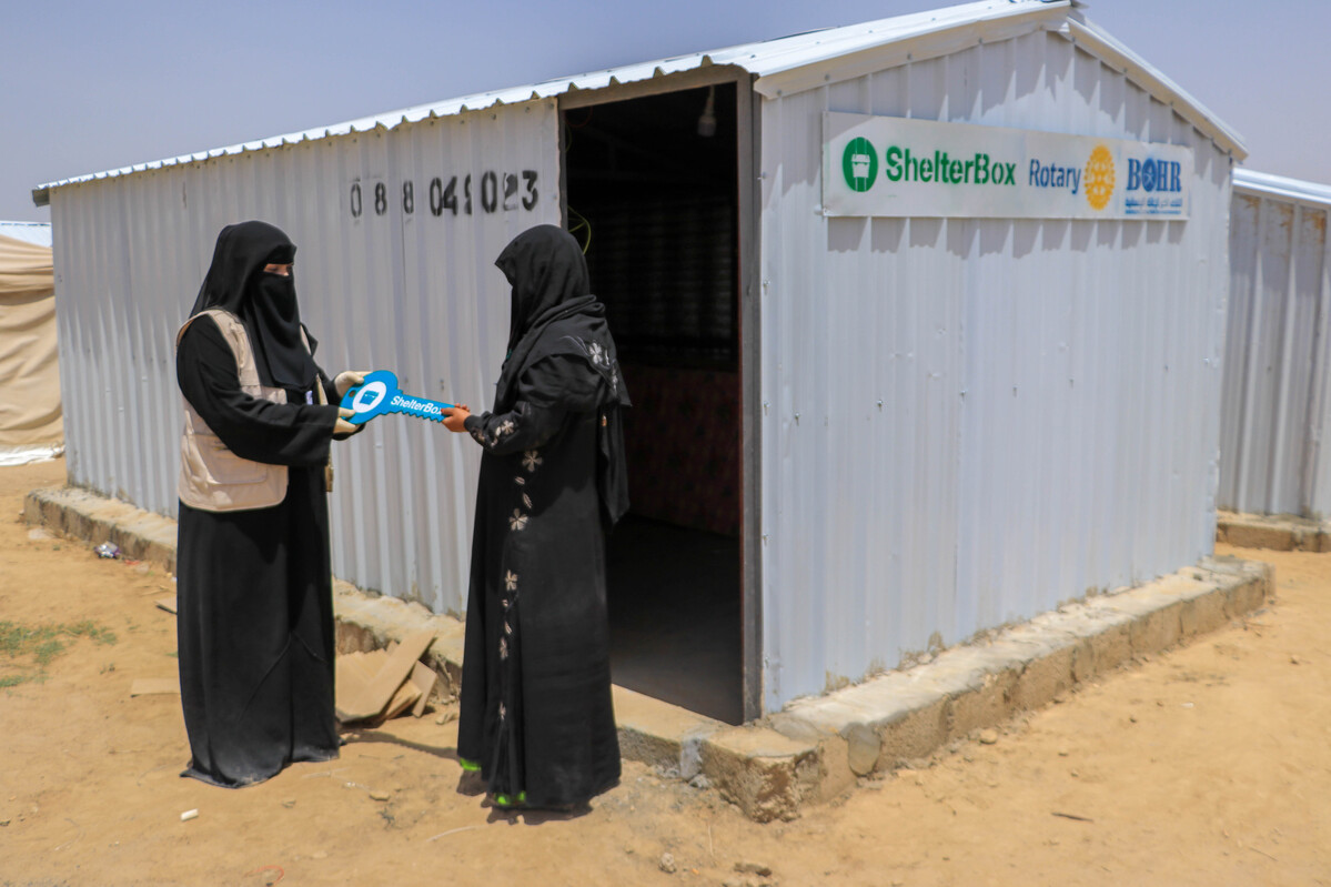 Woman handing a symbolic key to another woman outside of an iron net style shelter in Yemen