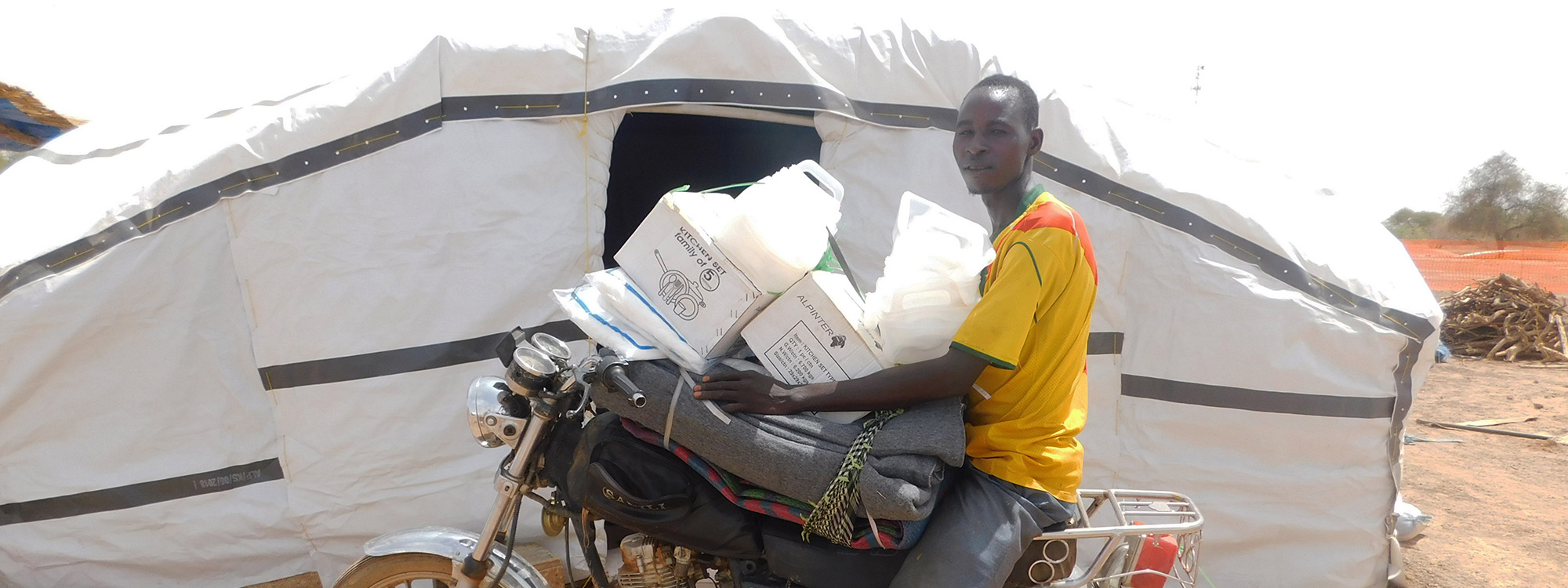 Man sitting on a motorbike holding aid items in front of a sahelian tent in Burkina Faso