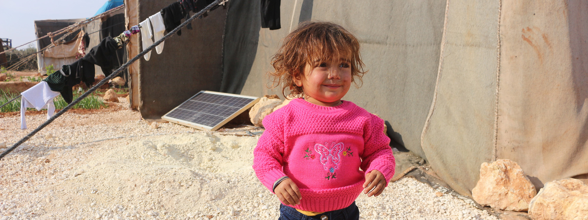 Child in a warm winter jumper next to tents in Syria