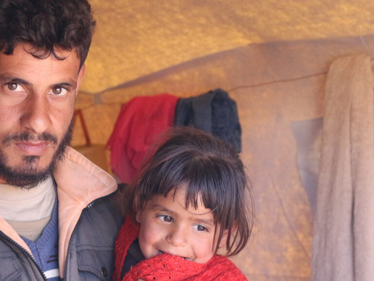 Man holding a child, both wearing winter clothes, inside a tent in Syria