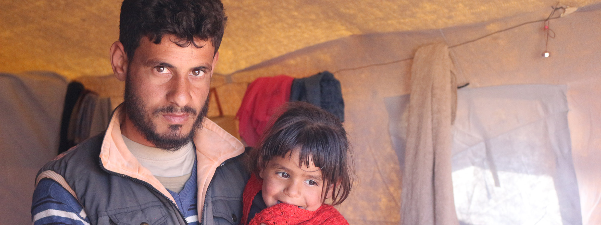 Man holding a child, both wearing winter clothes, inside a tent in Syria