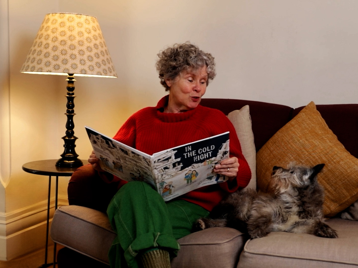 Imelda Staunton reading a book and looking at a dog sat next to her on the sofa