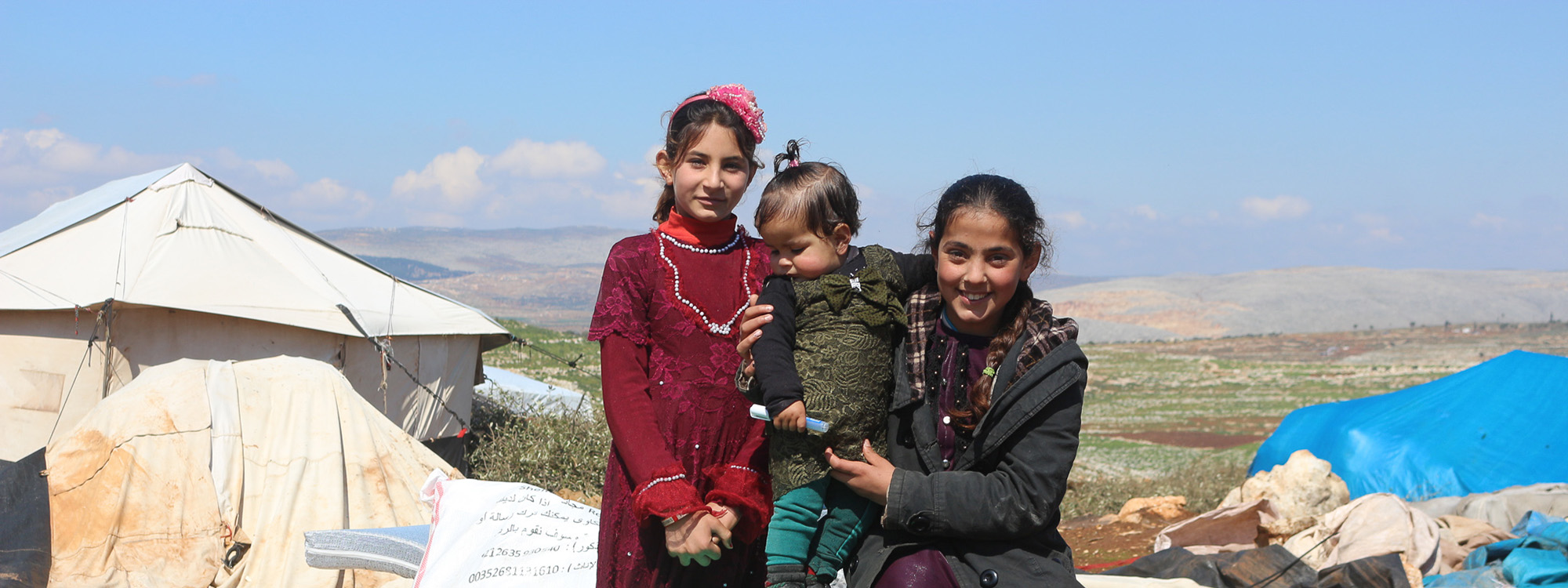 Three children in winter clothing standing outside a tent against the landscape in Syria