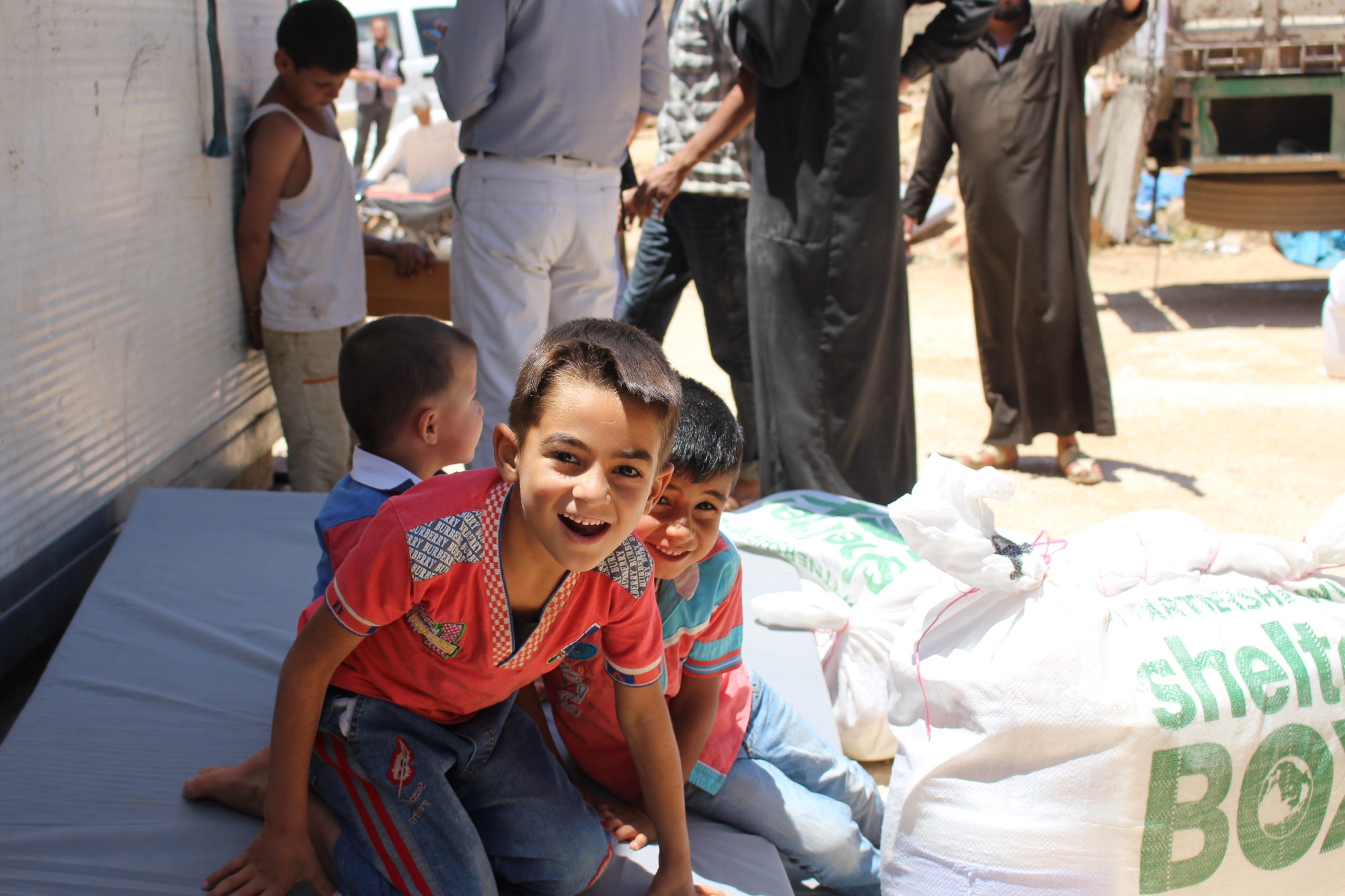 Children sitting on the floor smiling at the camera next to a bag of aid from ShelterBox and ReliefAid
