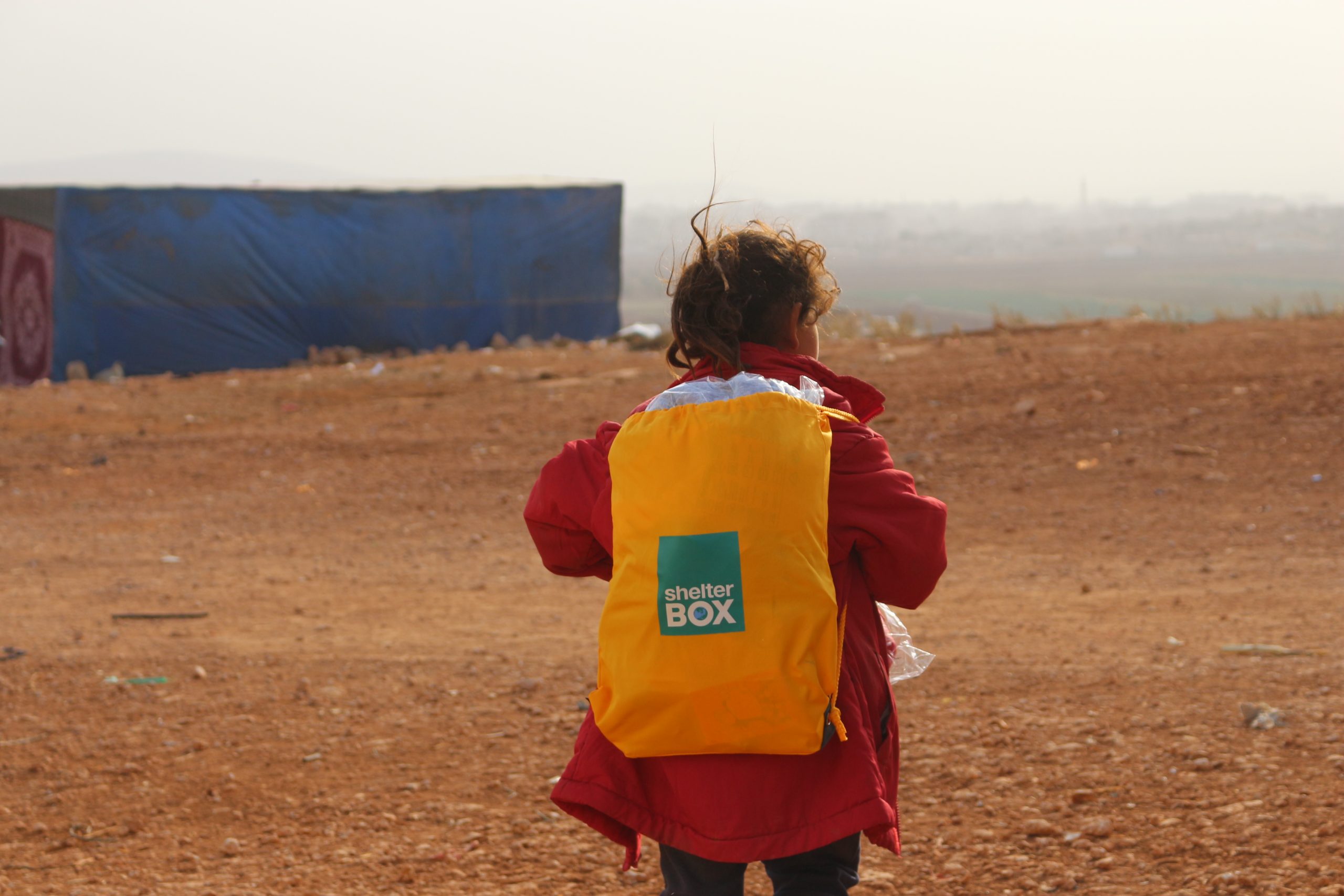 Child with their back to the camera, carrying a yellow backpack with the ShelterBox logo on it in Syria
