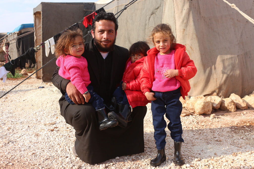 A father crouches in front of a tent, with his arms around his three young daughters, who are wearing warm coats, jumpers, jeans and waterproof boots.
