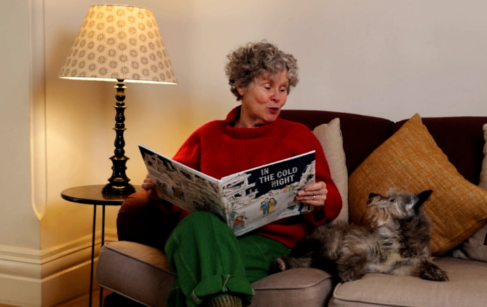 Imelda Staunton sit on a sofa with her dog reading from a children's booked called 'In The Cold Night'.