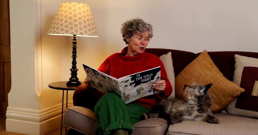 Imelda Staunton sit on a sofa with her dog reading from a children's booked called 'In The Cold Night'.