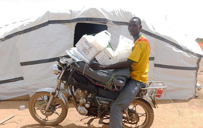 Man sitting on a motorbike holding aid in front of a sahelian tent in Burkina Faso