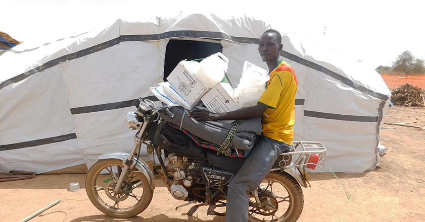 Man sitting on a motorbike holding aid in front of a sahelian tent in Burkina Faso