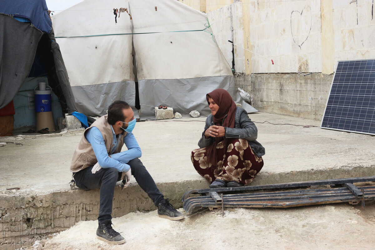 A ReliefAid worker wearing face mask and gloves talks to a woman during assessments at a camp in Idlib, Syria