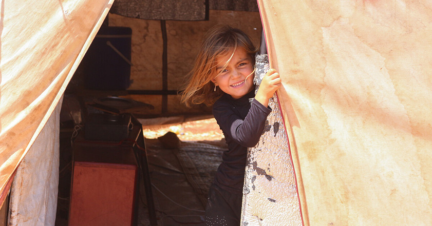 Child smiling while looking through the doorway of a tent in Syria