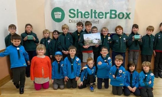 A group of children standing and kneeing in front of a ShelterBox banner