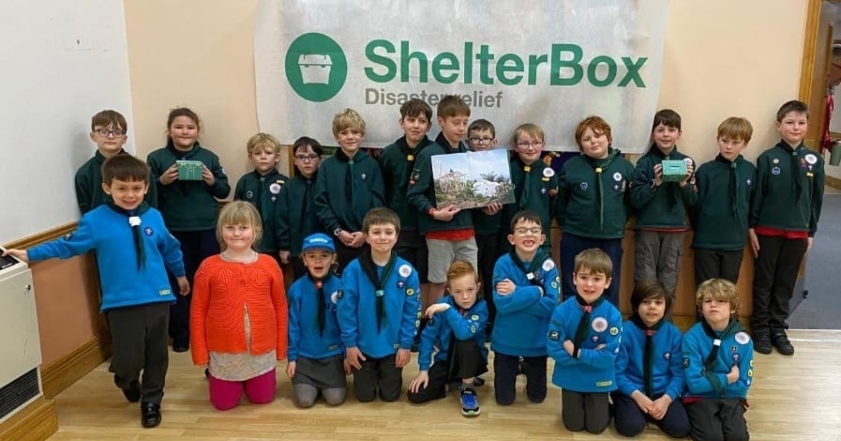 A group of children standing and kneeing in front of a ShelterBox banner