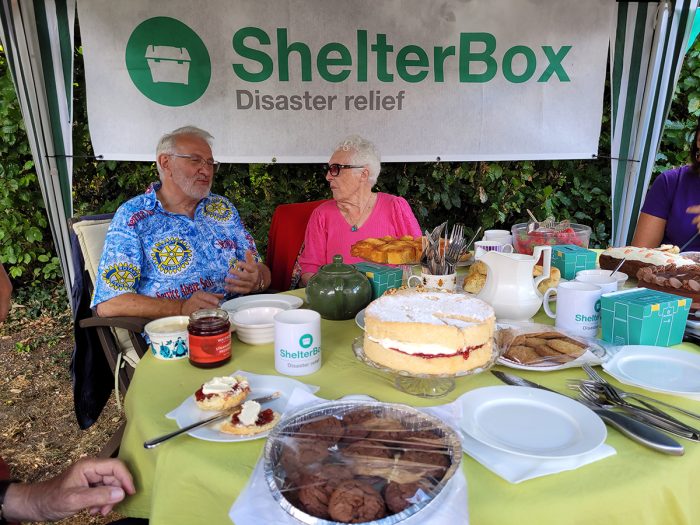 Two people in front of a ShelterBox banner and behind a table with cakes and drinks