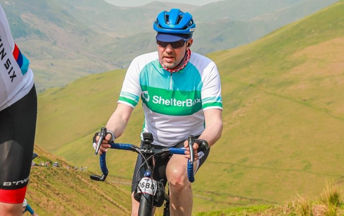 A man wearing a helmet and ShelterBox jersey rides a bike up hill with hills in the background