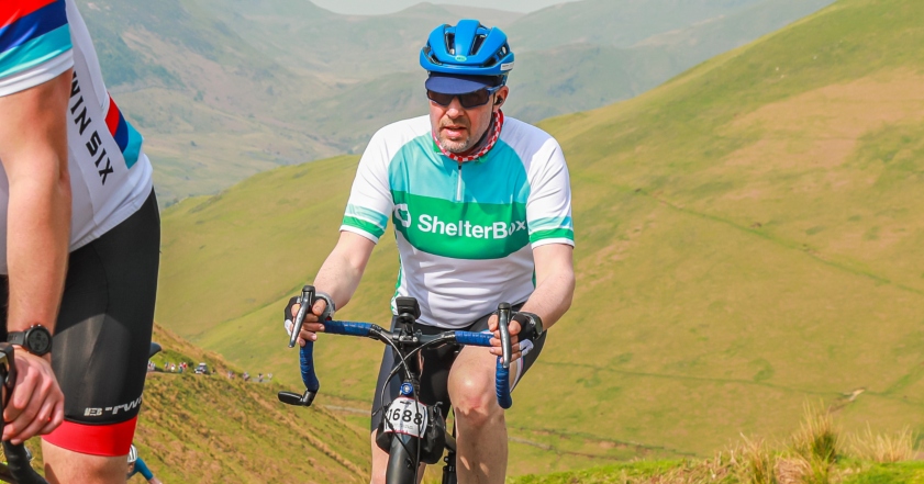 A man wearing a helmet and ShelterBox jersey rides a bike up hill with hills in the background