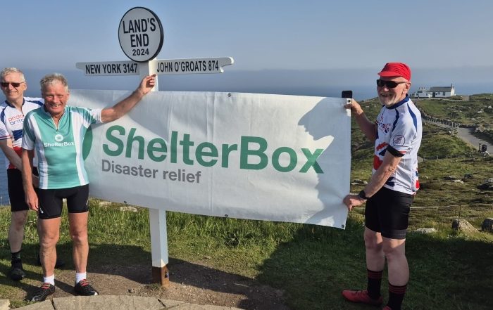 Three men holding a ShelterBox banner next to the sign post at Land's End