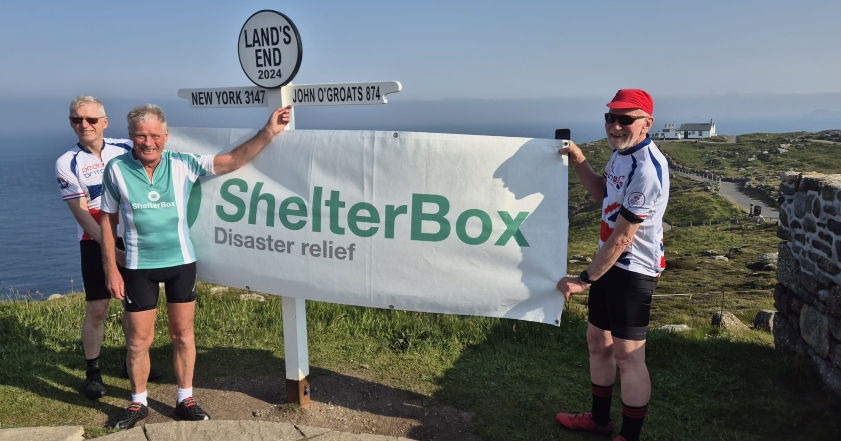 Three men holding a ShelterBox banner next to the sign post at Land's End