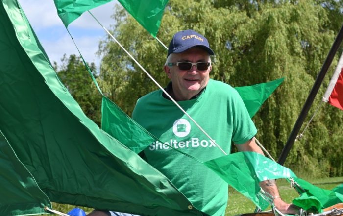 A man wearing a green ShelterBox t shirt stands inside a boat behind green sails.