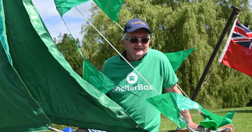 A man wearing a green ShelterBox t shirt stands inside a boat behind green sails.